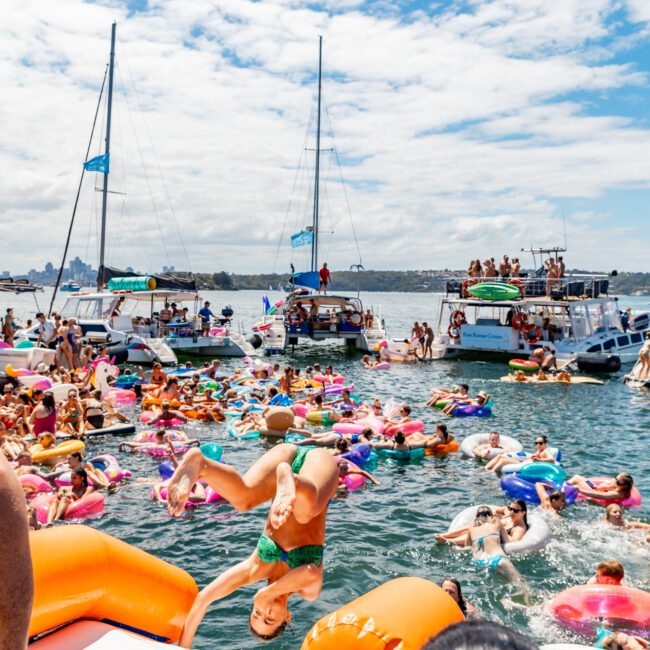 A vibrant scene at The Yacht Social Club Event Boat Charters, where people enjoy a sunny day on the water, floating on colorful inflatables. In the foreground, someone flips off an orange and white inflatable slide. Several boats are anchored in the background under a partly cloudy sky.