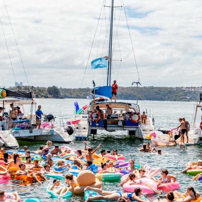 A colorful scene with many people floating on inflatable pool toys in the water, near several sailboats. The festive atmosphere is lively with participants enjoying the sunny weather. In the background, you can spot a cloudy sky and a distant shoreline, perfect for a day at The Yacht Social Club Sydney Boat Hire.