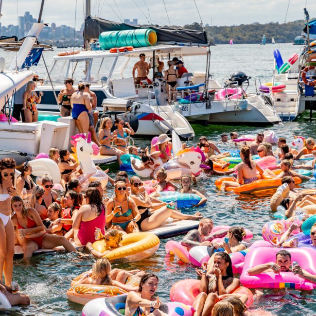 A vibrant scene shows a sunny day with a large group enjoying The Yacht Social Club. They are swimming and lounging on colorful inflatables. Several boats, including Luxury Yacht Rentals Sydney, are anchored nearby, with the city skyline visible in the distant background.