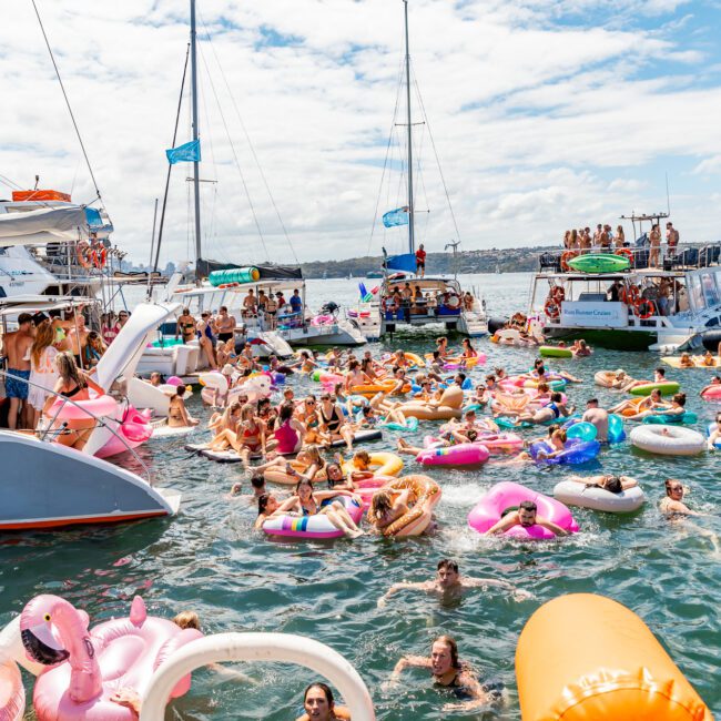 A lively scene of a boat party on a sunny day with many people floating on colorful pool inflatables in the water. Multiple boats are anchored nearby, with passengers both on board and in the water enjoying the festivities. The sky is partly cloudy. Welcome to Boat Parties Sydney The Yacht Social Club!