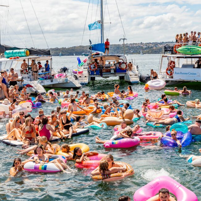 A lively gathering of people on colorful inflatable floats enjoying a sunny day on the water, surrounded by several anchored boats. The scene is festive, with guests of The Yacht Social Club Sydney Boat Hire swimming, relaxing, and socializing in a scenic bay area.