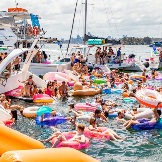 A lively scene of people enjoying a sunny day on the water with numerous colorful inflatable rafts and floaties. Boats are docked nearby, and everyone is having a good time, swimming and relaxing on various inflatable devices. The sky is partly cloudy, perfect for The Yacht Social Club Sydney Boat Hire experience.