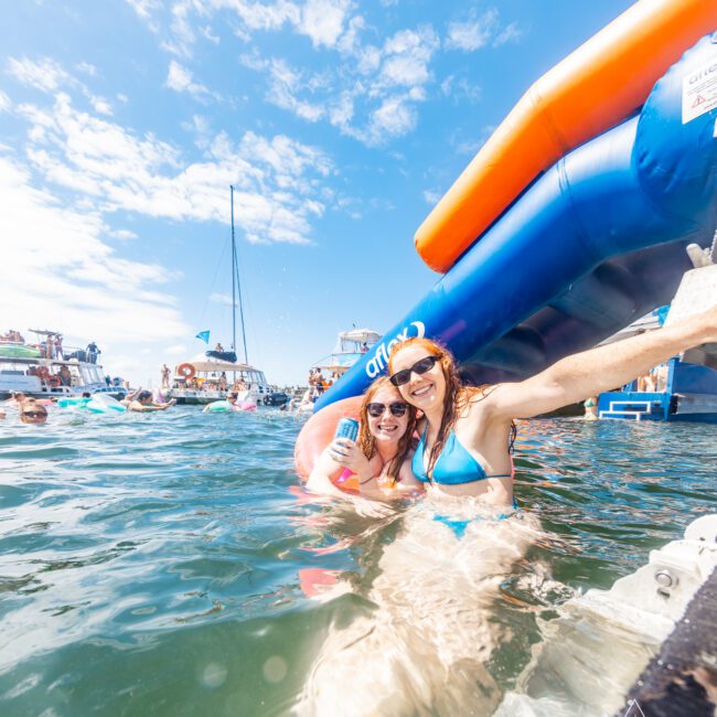 Two women in swimsuits smile for the camera while standing in the water next to a large inflatable slide, enjoying a sunny day with a clear blue sky. Other people, boats, and inflatables can be seen in the background. Consider joining The Yacht Social Club Sydney Boat Hire for an unforgettable experience on the water.