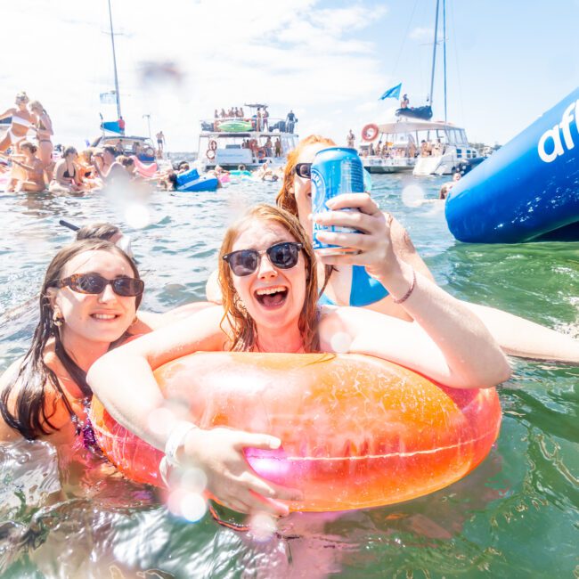 Two women enjoying a sunny day in the water, one holding a drink while sitting in an orange inflatable ring, both wearing sunglasses and smiling. Surrounding them are other people and boats from The Yacht Social Club Event Boat Charters, creating a lively and festive atmosphere.