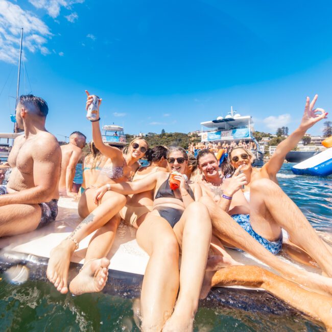 A group of people in swimwear are sitting on the back of a boat, holding drinks and posing for the camera. They are smiling and enjoying a sunny day on the water with other boats visible in the background. The scene is lively and vibrant with a clear blue sky overhead, perfect for Sydney Harbour Boat Hire The Yacht Social Club.