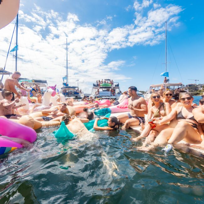 A lively group of people enjoying a sunny day on the water, surrounded by boats and colorful inflatable floaties. They are smiling, laughing, and swimming, with some seated on the edge of boats. The Yacht Social Club’s Sydney Boat Parties create a festive atmosphere under clear skies with scattered clouds.