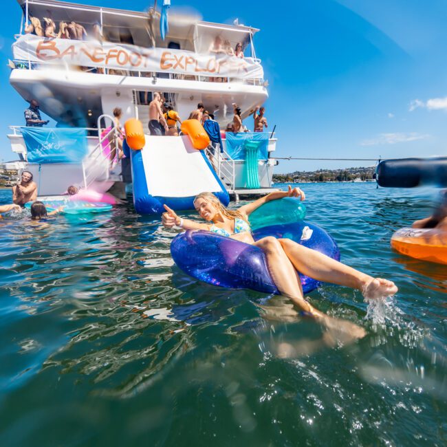 People are enjoying a sunny day on and around a yacht at The Yacht Social Club Event. Some are on the boat, while others float in the water on inflatable rings. A slide extends from the yacht into the water, with one person sliding down. The surroundings are calm, with clear blue water.