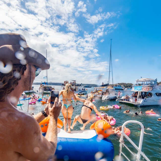 A person wearing a hat snaps a photo with a smartphone of two people in swimwear jumping off a boat into the water. Other boats and partygoers on colorful floatation devices are visible, enjoying The Yacht Social Club Event Boat Charters under a sunny, partly cloudy sky.