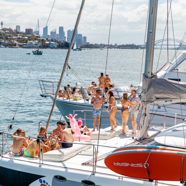 A group of people in swimwear are partying on two adjacent white sailboats on a sunny day, part of The Yacht Social Club event. One boat features a large inflatable pink flamingo. The city skyline is visible across the water, creating a lively and festive scene with clear blue skies above.