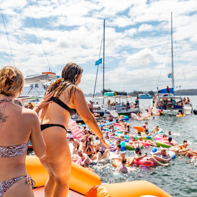 A lively scene of people enjoying a sunny day on the water. Dozens of colorful inflatable floats and boats are spread across the sea. Toward the foreground, individuals in swimsuits prepare to jump in from an inflatable platform at The Yacht Social Club Event Boat Charters.