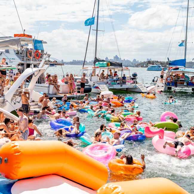 A lively scene of people enjoying a boat party on the water, courtesy of The Yacht Social Club Sydney Boat Hire. Numerous individuals float on colorful inflatable pool toys near several anchored boats. The weather is sunny with clear skies, and everyone appears to be having fun.