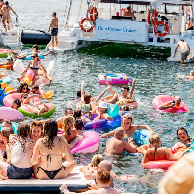 A vibrant party scene on a boat and in the water. People are enjoying time on colorful inflatable floats near a large boat named "Rum Runner Cruises." The backdrop includes other boats and a scenic coastline. Hosted by The Yacht Social Club, the atmosphere is lively and festive.