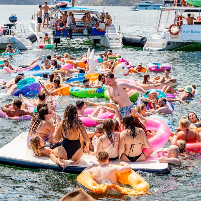 A lively scene of people enjoying a sunny day on a lake. Numerous individuals are floating on colorful inflatables, while a few swim and others sit on a platform in the water. Several boats are anchored nearby, reminiscent of Sydney Harbour Boat Hire The Yacht Social Club. The atmosphere is festive and vibrant.