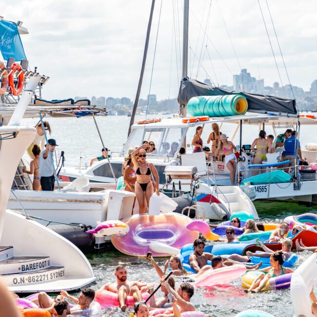 A vibrant scene on a sunny day with numerous people on inflatable floats enjoying a boat party on the water. Several boats are anchored nearby, including luxury yacht rentals from The Yacht Social Club. People are laughing, swimming, and having fun. The city skyline is visible in the background.