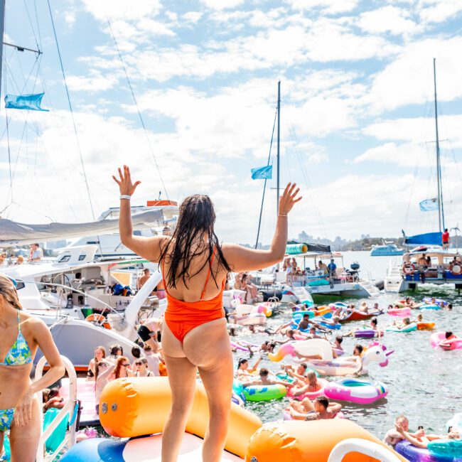 A person in a red swimsuit stands on an inflatable platform on a boat, arms raised high. The Yacht Social Club scene is lively, featuring numerous people on colorful rafts and floats surrounded by several boats. The sky is sunny with scattered clouds, perfect for a boat rental and parties Sydney experience.