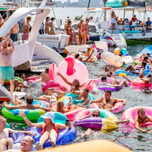 A lively scene with numerous people enjoying time on the water at The Yacht Social Club Event. Many are in colorful inflatable floaties, and several boats from Luxury Yacht Rentals Sydney are docked in the background. The sun is shining, and the atmosphere appears to be festive and social.