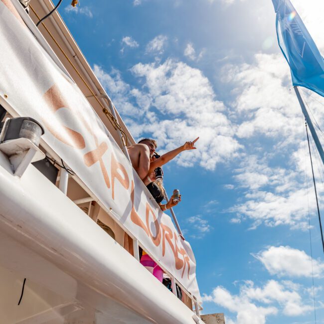 Two people stand on a boat's deck, one pointing into the distance and the other holding a cup. A large banner reading "EXPLORE" hangs on the railing. The sky is bright blue with scattered clouds. The scene conveys a sense of adventure and enjoyment, perfect for Sydney Harbour Boat Hire The Yacht Social Club.