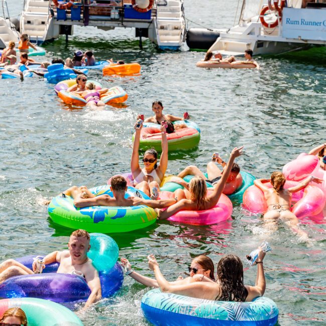 A lively scene of people enjoying a sunny day on the water, floating on colorful inflatable tubes. In the background, luxury yacht rentals from The Yacht Social Club Sydney Boat Hire are anchored. Many people are waving, holding drinks, and having fun in the water under the bright sky.