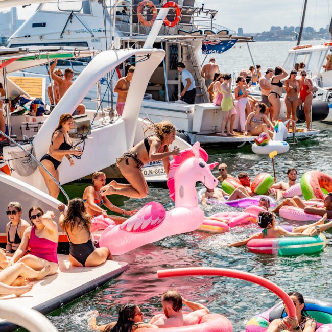 A lively scene on the water with people enjoying a boat party on a sunny day. Numerous people swim and float on colorful inflatable rafts, including a pink flamingo. Boats are docked nearby, with some partygoers dancing on the decks. Experience this at The Yacht Social Club Sydney Boat Hire for an unforgettable day.