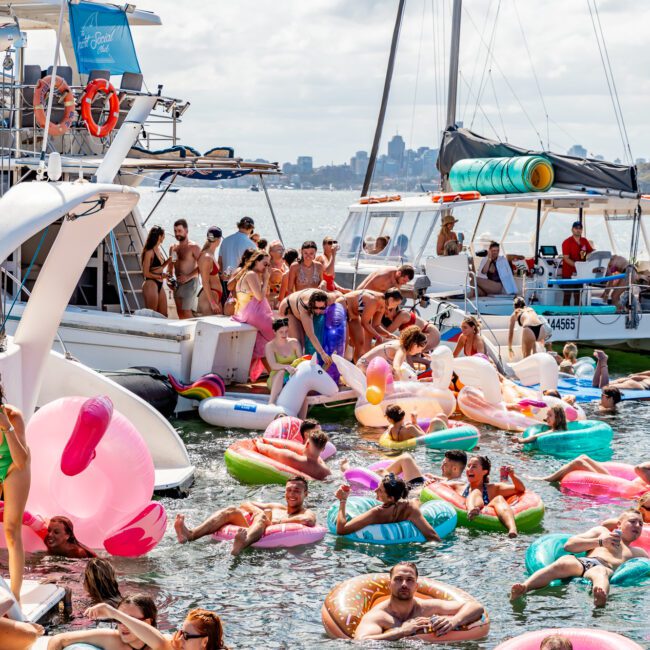 A vibrant scene of a boat party with numerous people enjoying the water on inflatable floaties. Various types and colors of floaties, boats, and a sailboat can be seen. The participants with The Yacht Social Club Sydney Boat Hire appear to be having a good time under a sunny sky with city buildings in the background.