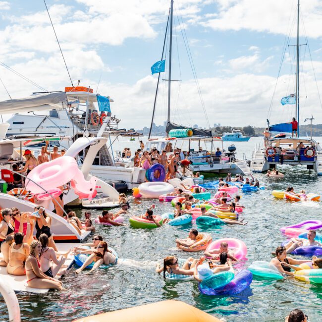 A lively scene of people enjoying a boat party on a sunny day. The water is filled with colorful inflatable floaties of various shapes and sizes, including a unicorn. Boats and yachts from Boat Rental and Parties Sydney The Yacht Social Club are docked nearby, with the shore visible in the background.