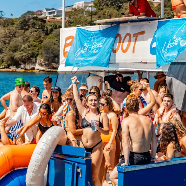 A group of people in swimwear are gathered on a boat with "BOOT" visible in the background. One woman in a black bikini stands on the edge, raising her hand and holding a cup. An inflatable slide leads to the water. The scene is sunny and festive, perfect for Boat Parties Sydney The Yacht Social Club.