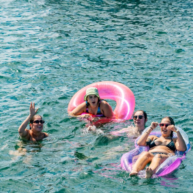 Four people enjoying time in the water near a boat. One person is using a pink pool float, another is on a purple float, and two others are swimming. One swimmer is making a peace sign, while another gestures animatedly. The Yacht Social Club Sydney Boat Hire offers the perfect backdrop with clear and calm waters.