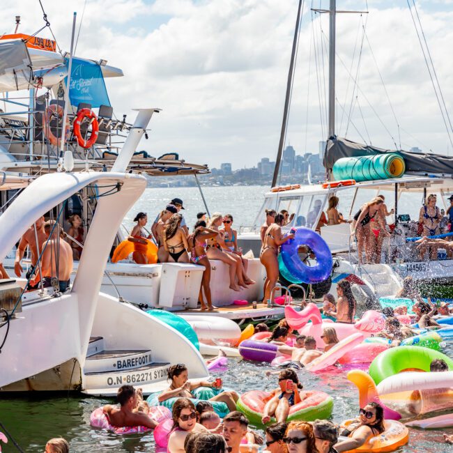 A lively scene with numerous people enjoying The Yacht Social Club event. Some are on boats, while others float in the water on colorful inflatables. The backdrop features the city skyline under a partly cloudy sky, making Boat Parties Sydney The Yacht Social Club truly memorable.