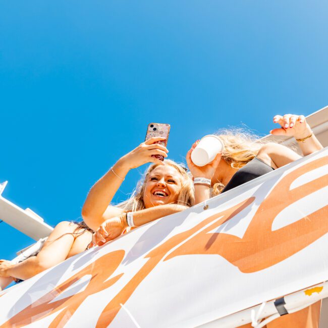 People enjoying a sunny day outdoors, taking selfies and holding drinks on an upper level or balcony, with clear blue skies above them. It's like a scene from The Yacht Social Club Sydney Boat Hire, perfect for boat rental and parties.