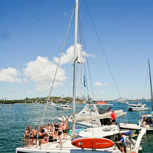 A lively group of people are celebrating on a white catamaran named "Barefoot," anchored in a picturesque harbor amidst several other boats. The sunny weather, with just a few clouds, perfectly highlights the charming shore lined with buildings and trees in the background.