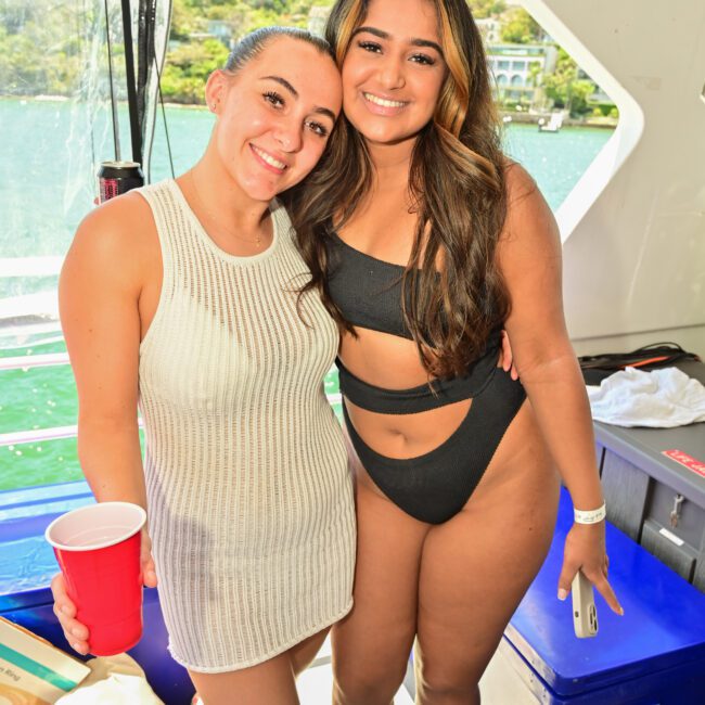 Two women pose side by side on a boat. The woman on the left is wearing a white sleeveless dress and holding a red cup, while the woman on the right is dressed in a black bikini. Both are smiling joyfully, with a scenic view of water and trees enhancing the serene backdrop.