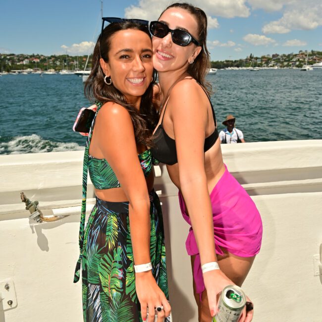 Two women are smiling and posing on a boat deck with the ocean and a coastal landscape in the background. One is wearing a tropical-patterned dress, while the other is sporting sunglasses with a black top and pink skirt. Both are enjoying canned beverages.