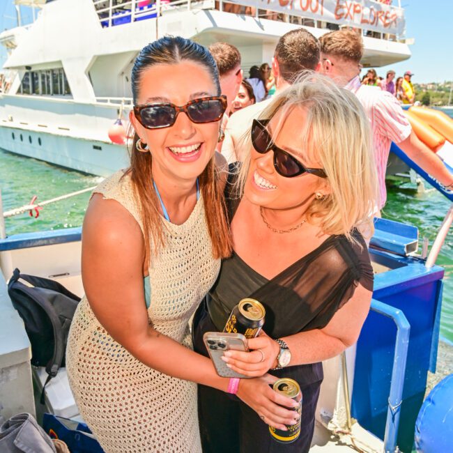 Two women smiling and holding drinks on the boat "Barefoot Explorer," surrounded by more people in the background. One woman wears a light-colored crochet dress, while the other is in a black outfit. The boat has guests on the upper deck, creating a festive and sunny atmosphere.