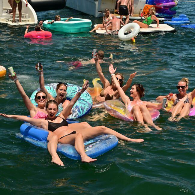 A group of people relax on colorful inflatables, holding drinks and smiling in a large body of water. Behind them is a boat with more people on board. The atmosphere is lively, with a blue sky and distant shoreline visible. A logo reads "The Yacht Social Club", epitomizing luxury and fun on the water.