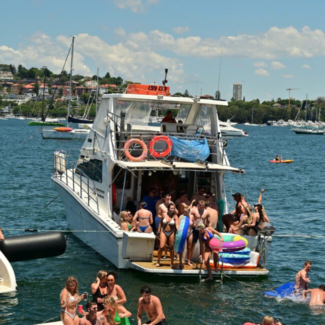 A dynamic scene of people enjoying a sunny day on a large boat and in the surrounding water. Many are on pool floats, swimming, or lounging on floating platforms. The boat is anchored near a picturesque coastline with various other boats visible in the background, creating a lively maritime atmosphere.