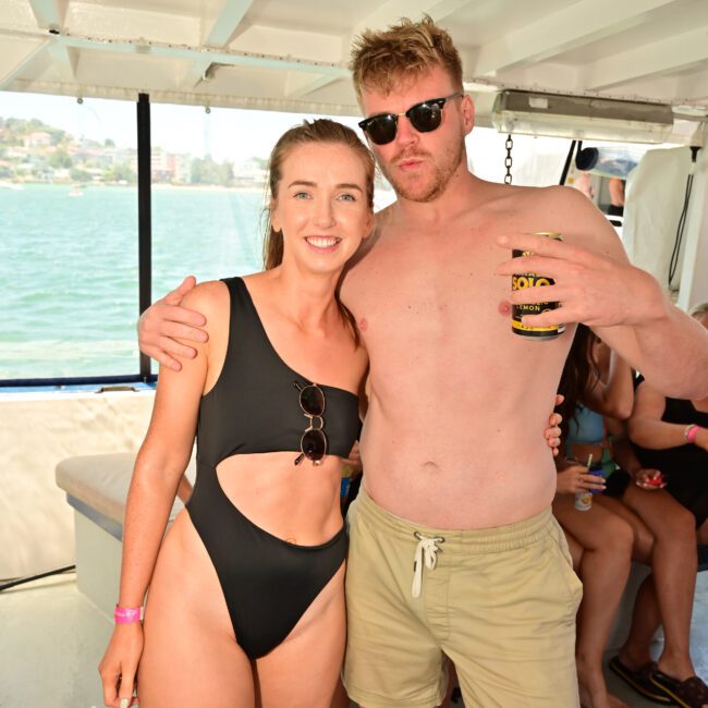 A man and woman pose together on a boat. The woman is wearing a black swimsuit and the man is shirtless with khaki shorts, holding a drink. Both are smiling at the camera, enjoying their Yacht Social. Other people are visible in the background.