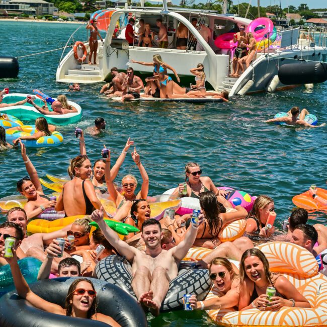 A lively scene of a large group of people enjoying a boat party on the water. Many are on colorful inflatable floats, holding drinks, and smiling at the camera. Behind them is a sailboat with additional partygoers holding parasols. The atmosphere is festive and joyful.