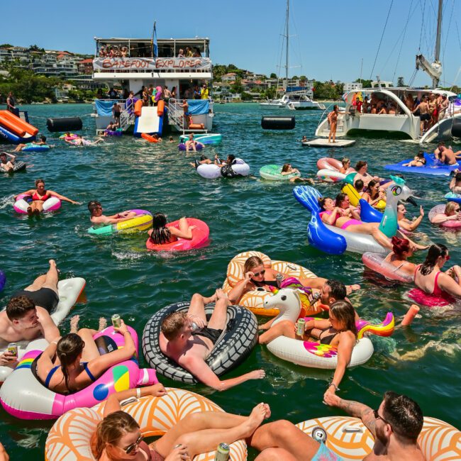 A lively scene on a sunny day with numerous people floating on colorful inflatable rafts and tubes in a lake. Two large boats with more people onboard are anchored in the background, and lush greenery is visible along the shore.