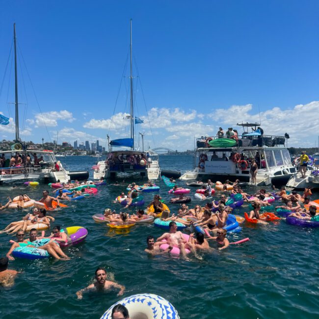 A lively scene of people enjoying a sunny day in the water, surrounded by several boats. Many are on colorful inflatable floats, and some are swimming. The sky is clear with a few clouds, and a sense of fun and relaxation is evident. Text reads "Yacht Social Club - Nautical Bliss.