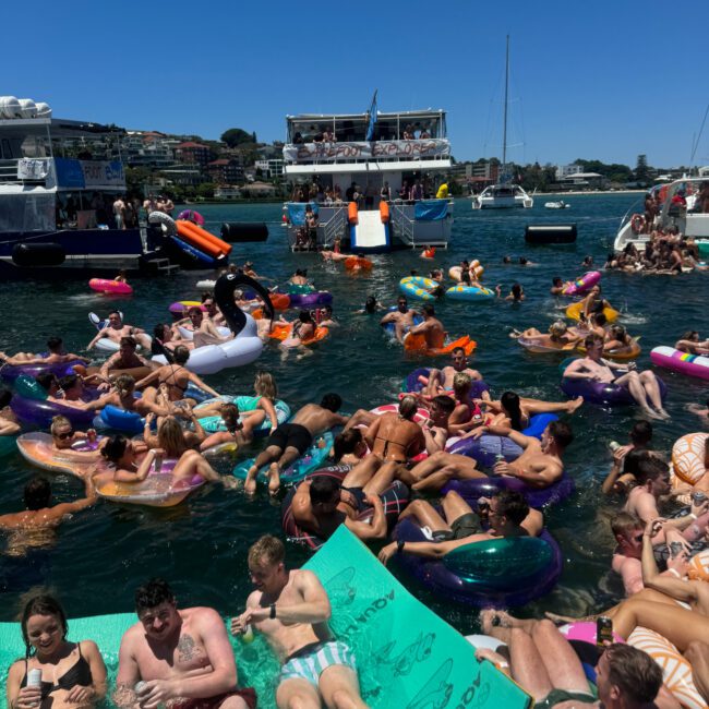 A lively scene of people enjoying a sunny day on a lake. Numerous individuals are floating on colorful inflatable rafts, including a pink flamingo and glittering unicorn. Boats are anchored nearby, with some people gathered on their decks. The sky is clear and blue.