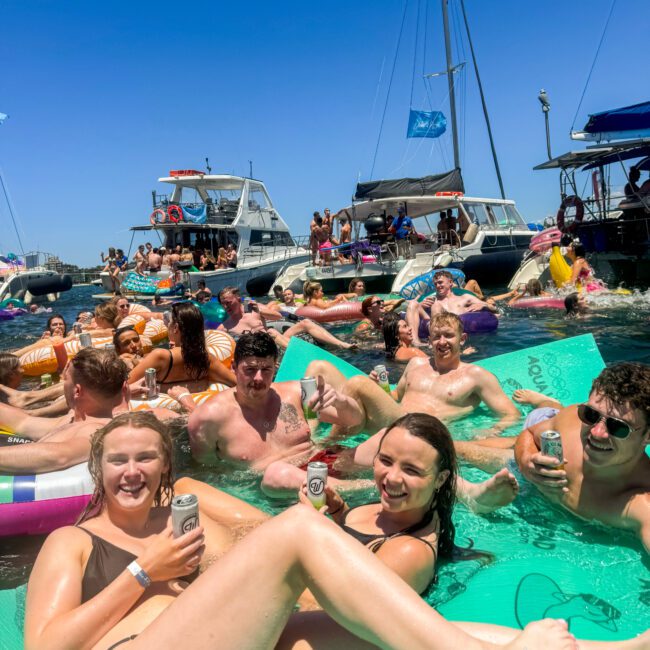 A lively group of people enjoying a sunny day on the water, floating on inflatables and large mats, holding drinks, and smiling. Boats are anchored nearby with more people visible on them. The atmosphere is festive and relaxed as laughter fills the air.