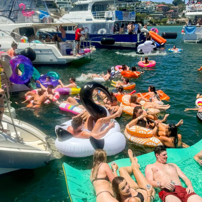 A lively scene of people enjoying a sunny day on the water. Many are floating on colorful inflatable pool toys in various shapes. Boats are docked nearby, and some individuals are relaxing on a large floating mat while sipping drinks. The background shows waterfront houses and lush trees.