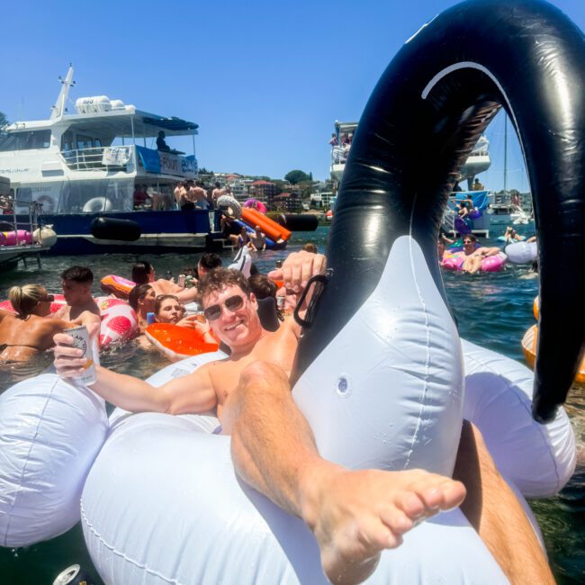 A man relaxes on a large inflatable swan float in the water, holding a drink. He is surrounded by other people in the water and nearby boats under a clear, sunny sky. Text in the bottom right corner reads "Yacht Social Club - Floating Paradise.