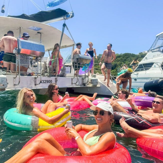 A lively scene of people enjoying themselves in a sunny outdoor setting. Several individuals float on vibrant inflatable rings in the water, holding drinks and smiling. Two boats are docked nearby, with more people socializing on the deck. Majestic trees can be seen in the background.