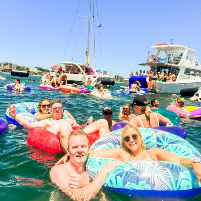 A group of people enjoying their time in the water, lounging on colorful inflatable rings. In the background, two boats and more people are socializing under a clear blue sky. The setting appears to be a lively social event or party on the water.