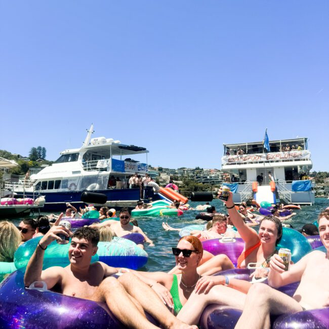 A lively group of young adults relax in colorful inflatable tubes on the water, surrounded by others enjoying the sunny day near two docked yachts. Some people are taking selfies with their smartphones, and others are holding drinks. The scene is vibrant and festive.