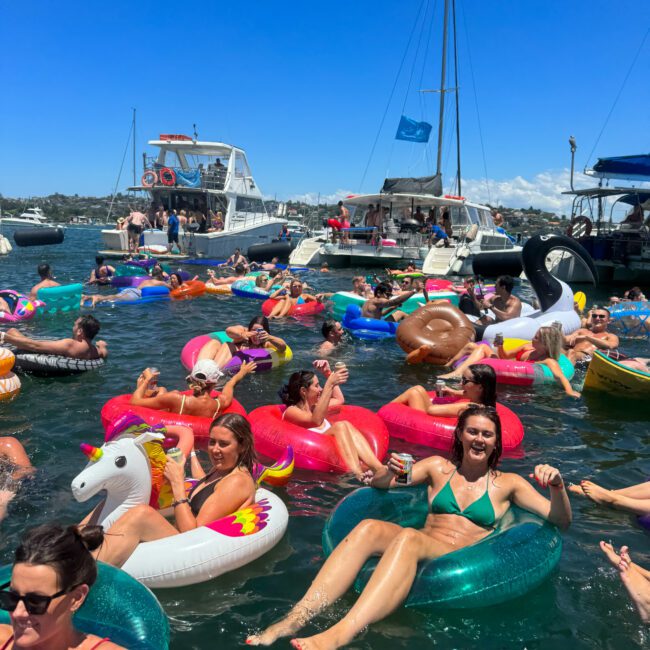 A lively scene of people floating on colorful inflatable rings in a sunny bay. Paddleboarders navigate gracefully nearby, and the water is filled with smiling individuals enjoying the warm, clear day. A blue sky overhead completes the vibrant atmosphere.