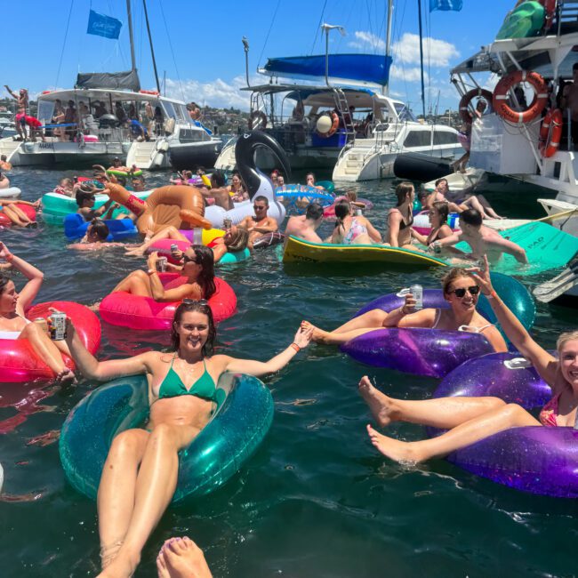 People are enjoying a sunny day on the water, relaxing on colorful inflatable pool floats with drinks in hand, surrounded by boats. The atmosphere is festive and lively, with a mix of laughter, vibrant summer outfits, and a variety of unique inflatables.