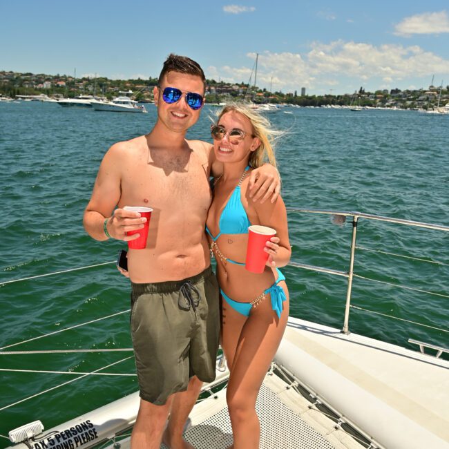 A man and a woman stand arm-in-arm on a boat deck, both holding red cups. The man wears sunglasses and swim trunks, while the woman is in a blue bikini and sunglasses. The water and several boats are visible in the background under a clear sky.