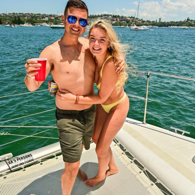 A smiling shirtless man in green shorts and sunglasses holds a red plastic cup. A woman in a yellow swimsuit stands next to him, also smiling. They are on the deck of a boat, with water and a captivating cityscape in the background.